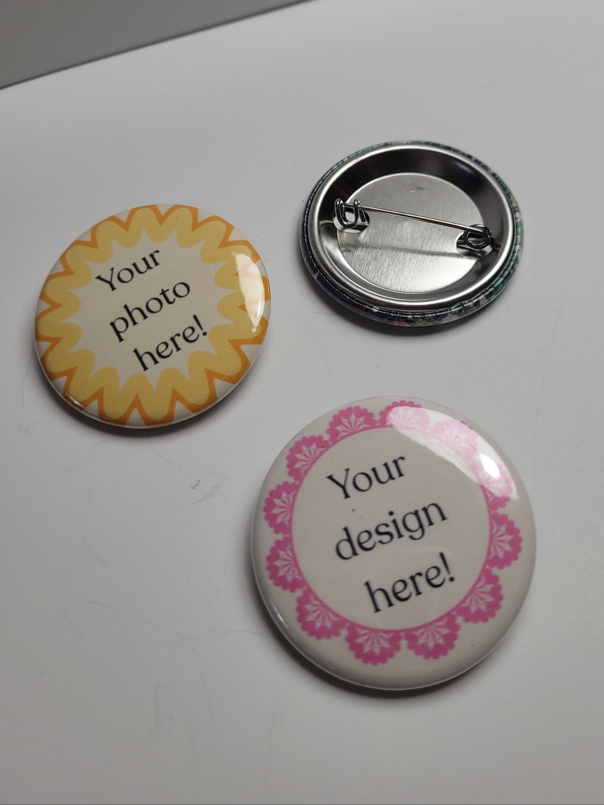 Three customizable round pinback buttons on a light gray surface. One button has a yellow sunburst border with the text 'Your photo here!', one has a silver metal back, and one has a pink floral border with the text 'Your design here!'.